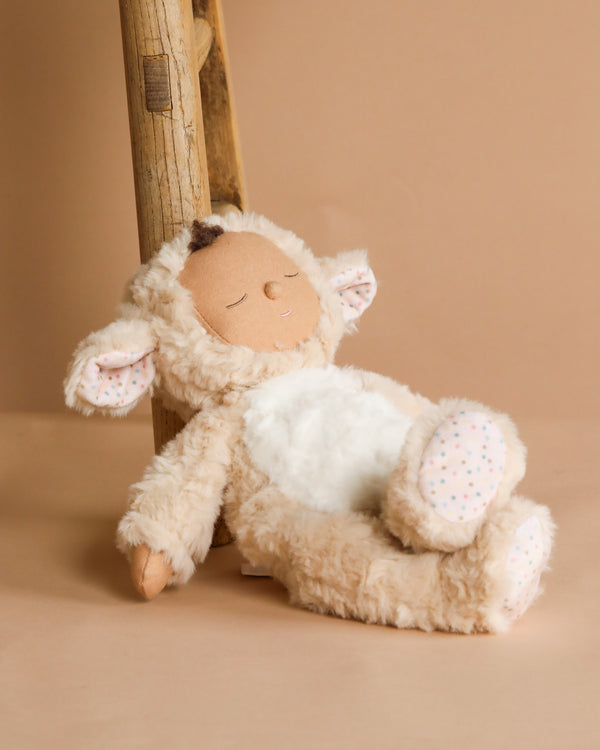 A Cozy Dinkums plush toy, the Olli Ella Cozy Dinkum Doll - Lamby Picket, leans against a wooden ladder with soft beige fur, patterned inner ears and paws, and a peaceful embroidered face. Set against a warm neutral background, it's the perfect Easter surprise plush companion.
