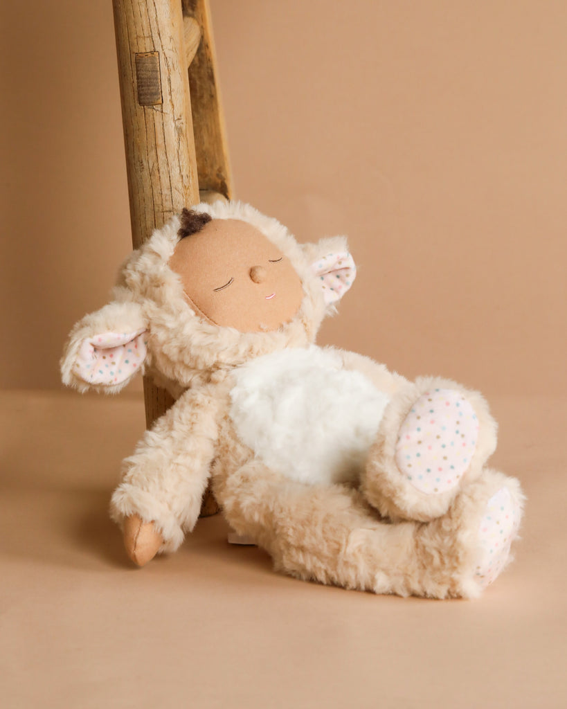 A Cozy Dinkums plush toy, the Olli Ella Cozy Dinkum Doll - Lamby Picket, leans against a wooden ladder with soft beige fur, patterned inner ears and paws, and a peaceful embroidered face. Set against a warm neutral background, it's the perfect Easter surprise plush companion.