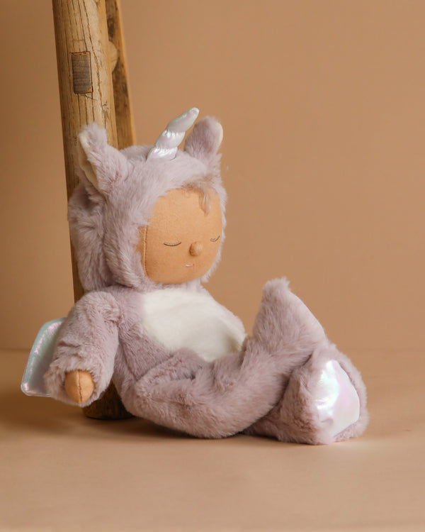 The Olli Ella Cozy Dinkum Doll Unicorn - Sweetpea is your ideal whimsical companion, dressed in a fuzzy unicorn costume with a horn and soft lavender fur, peacefully leaning against a wooden ladder on a warm beige background.