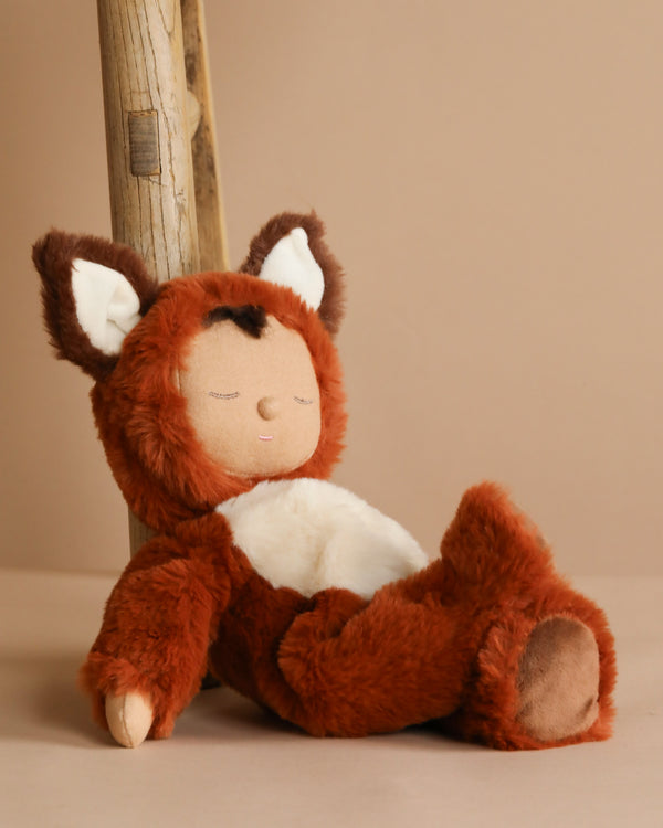 The Olli Ella Cozy Dinkum Doll - Fox, dressed in a brown and white costume with fox-like ears, leans against a wooden post on a beige background, exuding charm with its peaceful expression and closed eyes.