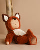 The Olli Ella Cozy Dinkum Doll - Fox, dressed in a brown and white costume with fox-like ears, leans against a wooden post on a beige background, exuding charm with its peaceful expression and closed eyes.