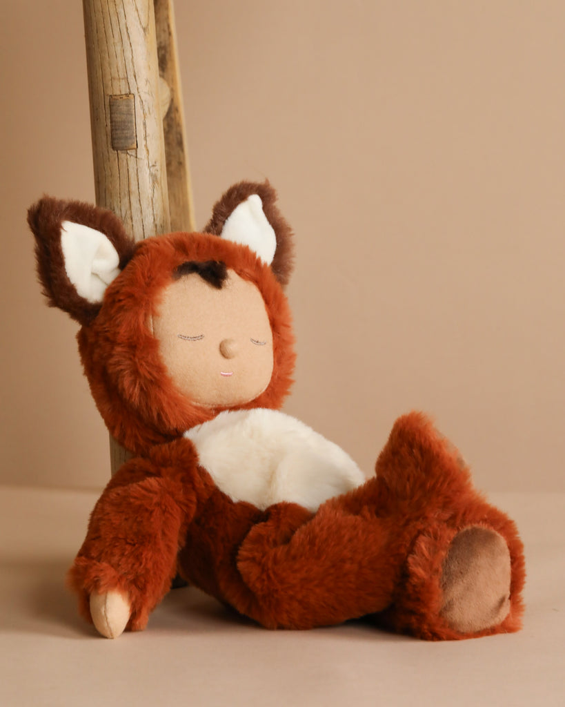 The Olli Ella Cozy Dinkum Doll - Fox, dressed in a brown and white costume with fox-like ears, leans against a wooden post on a beige background, exuding charm with its peaceful expression and closed eyes.