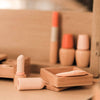 A close-up of minimalist makeup items from the Wooden Beauty Kit on a beige surface, featuring lipstick, compact, and foundation. The packaging in soft pastel shades enhances the warm, neutral aesthetic against the blurred background.
