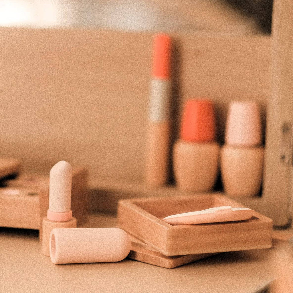 A close-up of minimalist makeup items from the Wooden Beauty Kit on a beige surface, featuring lipstick, compact, and foundation. The packaging in soft pastel shades enhances the warm, neutral aesthetic against the blurred background.