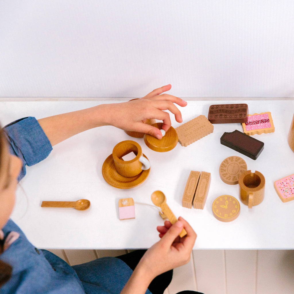 A child enjoys playtime with the Wooden Tea Set on a white table, arranging cups, saucers, and spoons with pastries, imagining an almond chai latte for a delightful afternoon of pretend tea time.