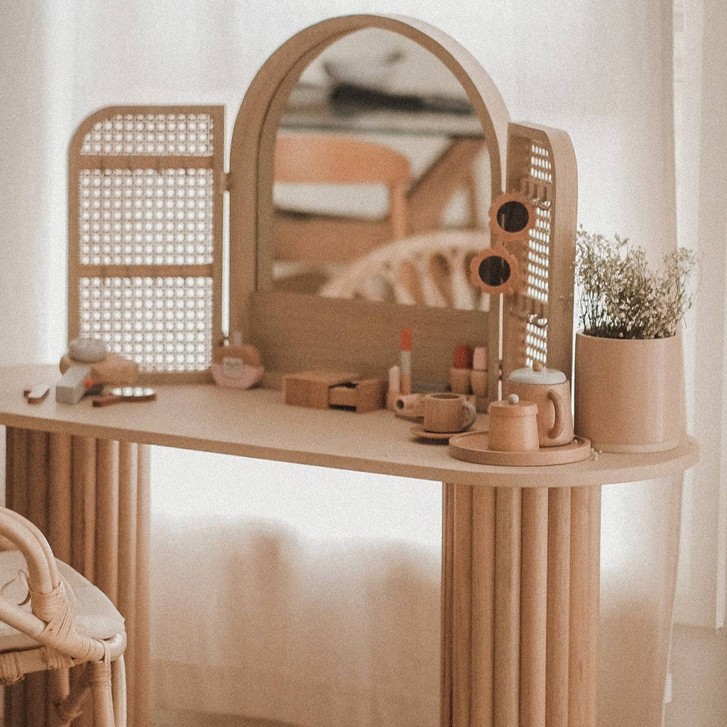 The Wooden Beauty Kit, a wooden vanity table with a round mirror and perforated side panels, painted in non-toxic hues, holds cosmetics, lipsticks, and a floral vase. It features makeup accessories in a softly lit room with wicker and natural elements.