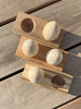 Three rectangular Wooden Egg Shakers with Stand are arranged diagonally on a light wood surface; two feature two smooth, egg-shaped knobs, while the third has two round, shallow depressions.