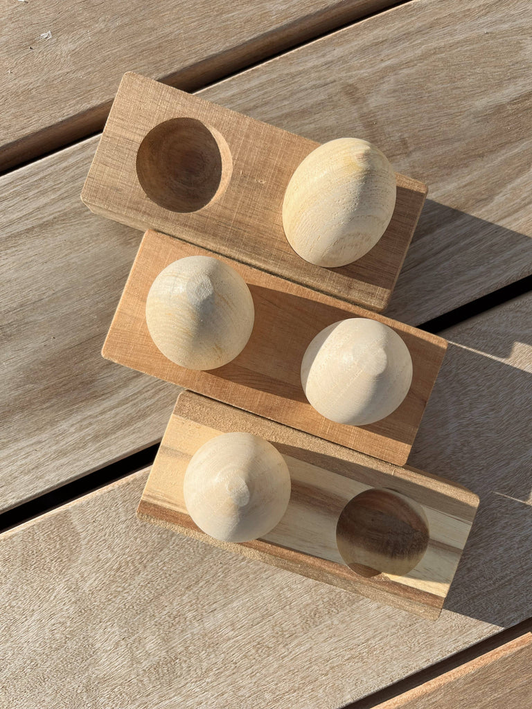 Three rectangular Wooden Egg Shakers with Stand are arranged diagonally on a light wood surface; two feature two smooth, egg-shaped knobs, while the third has two round, shallow depressions.