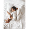 A young child dressed in white sleeps on a bed, cuddling the Senger Naturwelt Cuddly Animal - White Seal, with another white seal warming pillow nearby. The all-white bedding and plush toys create a soft, cozy scene.