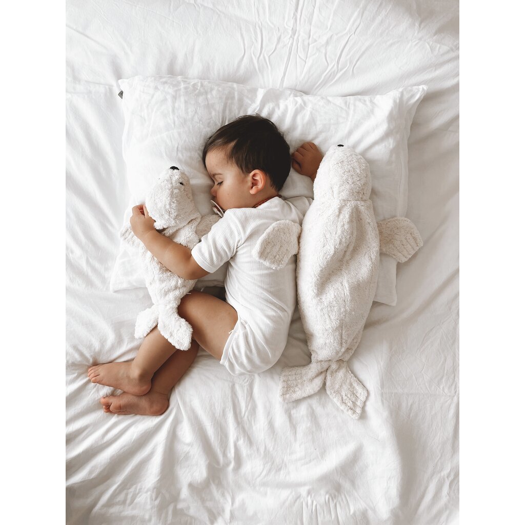 A young child dressed in white sleeps on a bed, cuddling the Senger Naturwelt Cuddly Animal - White Seal, with another white seal warming pillow nearby. The all-white bedding and plush toys create a soft, cozy scene.
