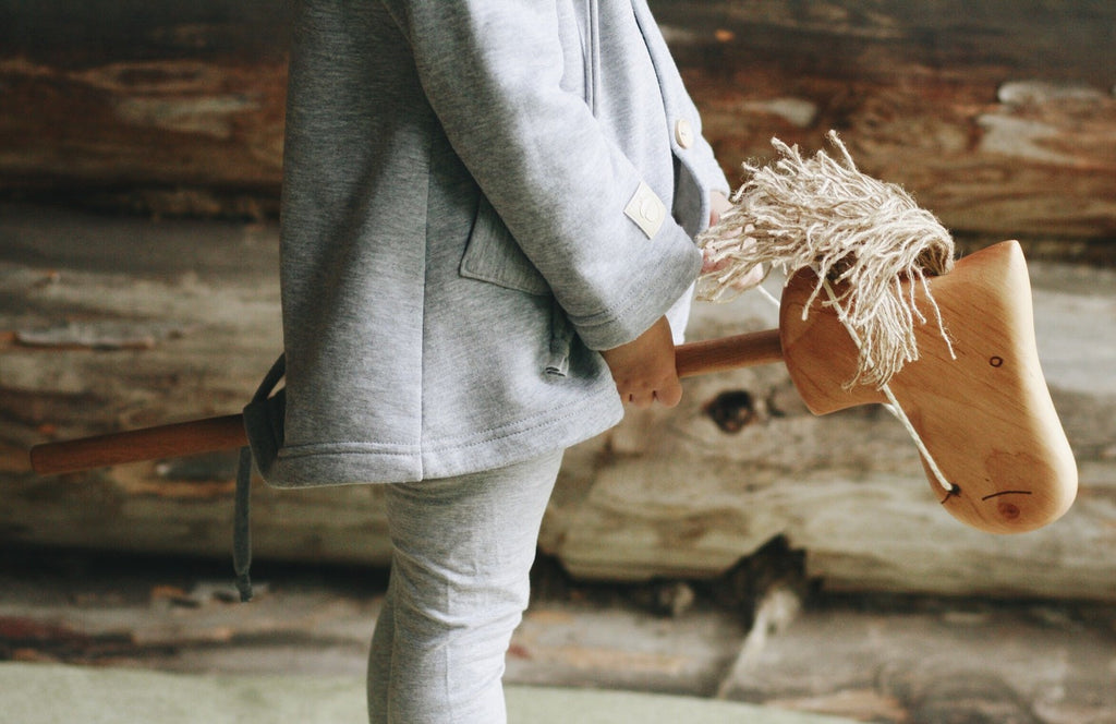 A child in grey holds a Handmade Wooden Riding Horse with a yarn mane, set against a backdrop of wooden logs for a rustic touch.
