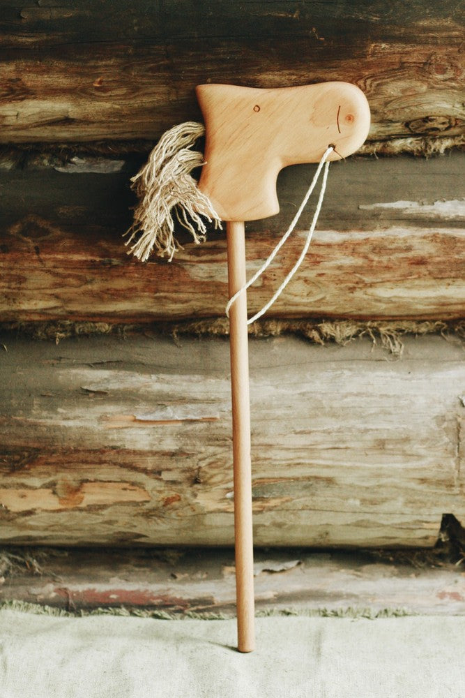 A Handmade Wooden Riding Horse, crafted from beech wood with a carved head and yarn mane, rests against a rustic log wall. Its simple design and twine reins exude charm and nostalgia.