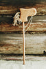 A Handmade Wooden Riding Horse, crafted from beech wood with a carved head and yarn mane, rests against a rustic log wall. Its simple design and twine reins exude charm and nostalgia.