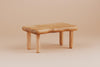A Maileg Dining Room Set small table, with a natural wood finish and smooth, rounded edges on four round legs, stands beside a Maileg Flower Rug against a beige background.
