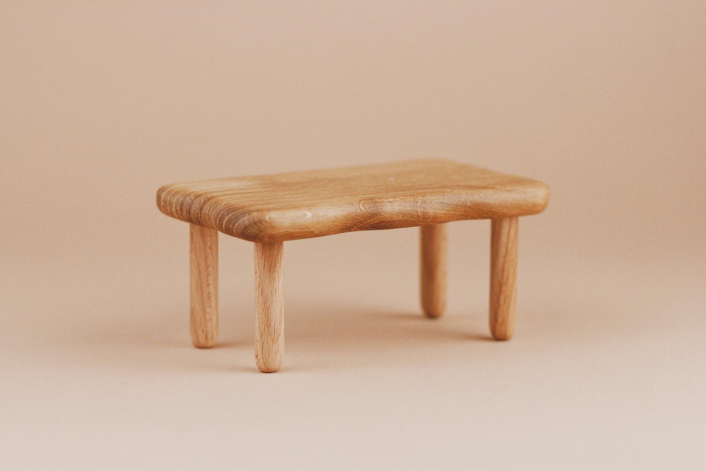 A Maileg Dining Room Set small table, with a natural wood finish and smooth, rounded edges on four round legs, stands beside a Maileg Flower Rug against a beige background.