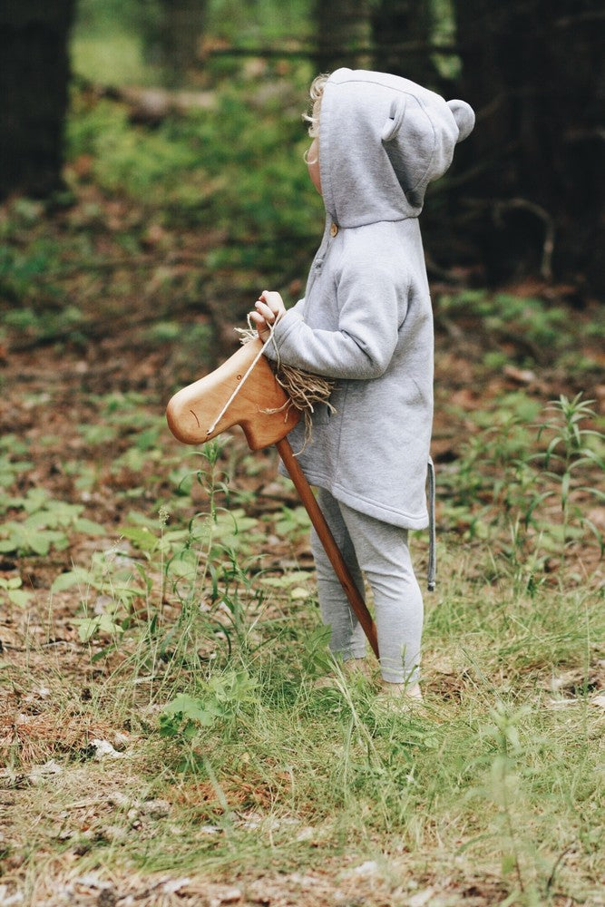 In a grassy, wooded area, a young child wearing a grey hoodie with bear-like ears playfully and exploratively clutches a Handmade Wooden Riding Horse, with their face turned away.