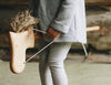 A child, dressed in gray, stands indoors on a light floor, holding a Handmade Wooden Riding Horse with a yarn mane.