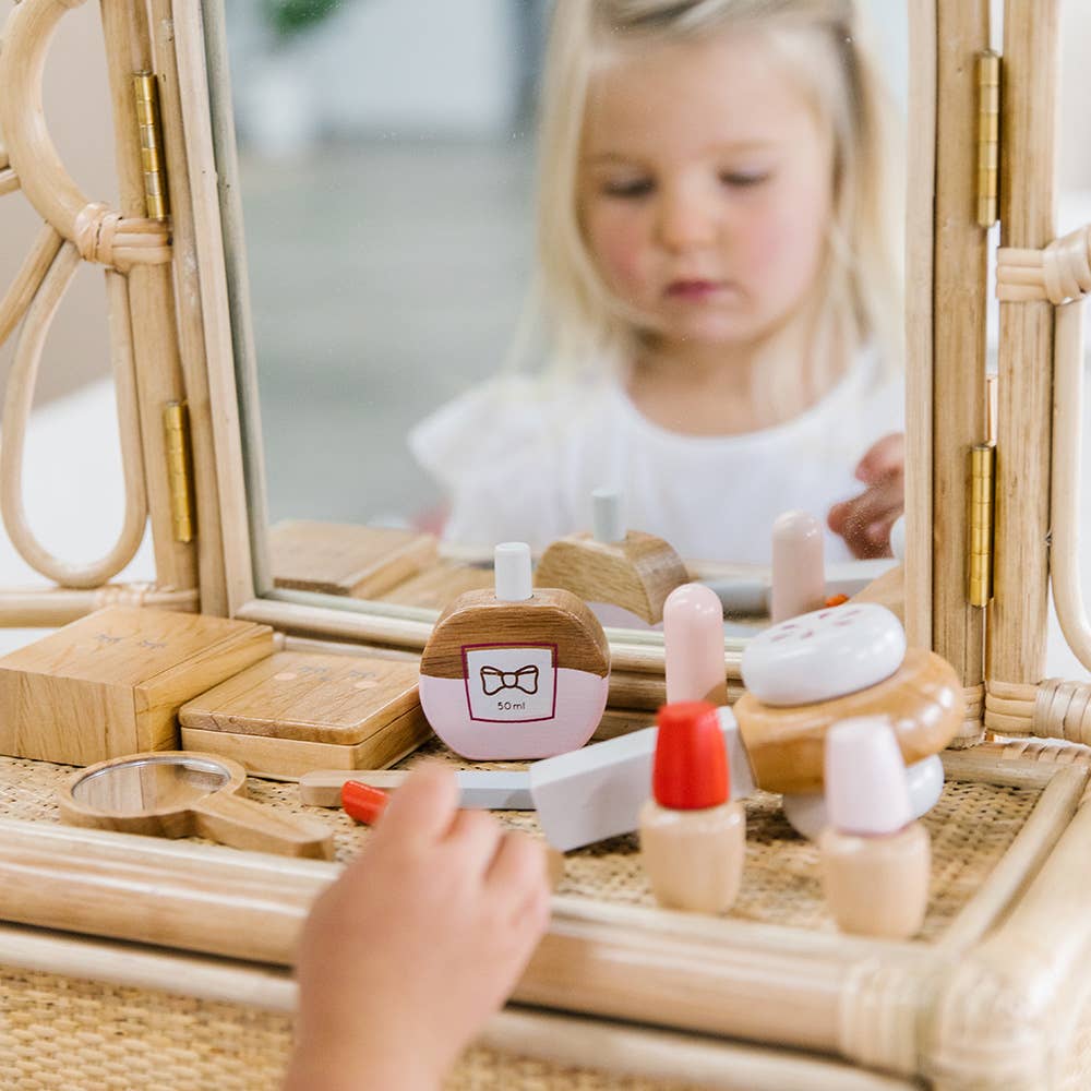 A young child is captivated by the Wooden Beauty Kit at a vanity, playing with toy makeup items like pretend lipstick and nail polish, all made from non-toxic paints on a woven surface. The scene beautifully captures the timeless joy of imaginative play.