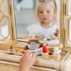 A young child is captivated by the Wooden Beauty Kit at a vanity, playing with toy makeup items like pretend lipstick and nail polish, all made from non-toxic paints on a woven surface. The scene beautifully captures the timeless joy of imaginative play.