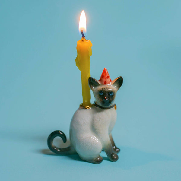 The Siamese Cat Cake Topper is a charming ceramic figurine with a red party hat and lit yellow candle, set against a serene light blue background. Perfect for cat-themed parties, it adds whimsy to any celebration.