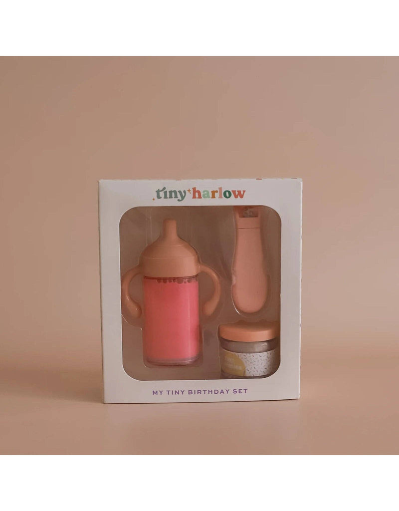 The "My tiny birthday set - Tiny Harlow" is a pink and peach-toned toy set with a magical sippy cup, toy spoon, and birthday cake jar, perfect for a doll's party. It's beautifully packaged in a box against a beige background.