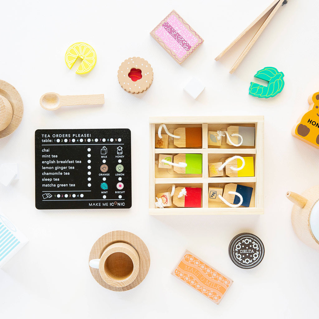 A flat lay of the Wooden Tea Set includes toy tea bags like organic Matcha Tea in a wooden box, a wooden cup, assorted toy pastries, and a "Tea Orders" board. This vibrant set enhances imagination on any play area's white surface.