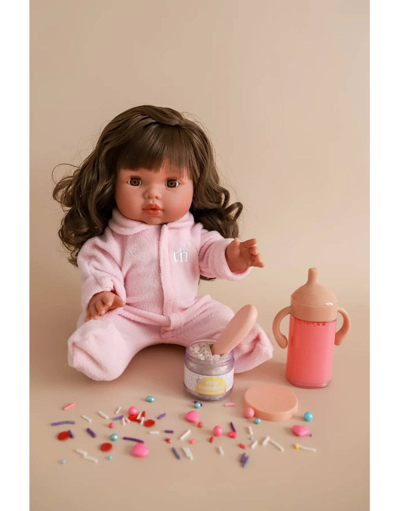 The "My Tiny Birthday Set" from Tiny Harlow features a long brown-haired doll in a pink onesie on a beige background. It includes colorful candies, a white cream jar like a birthday cake, and pink play accessories: spoon and handled baby bottle for realistic play.