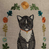 An embroidered black and white Tuxedo Cat with a red collar, surrounded by vibrant spring flowers and leaves on beige fabric, adorns the Coral & Tusk Kitten Cameo Pillow. Ships in one week.