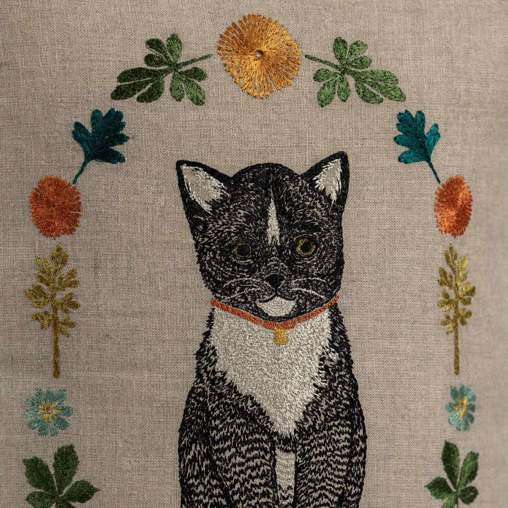 An embroidered black and white Tuxedo Cat with a red collar, surrounded by vibrant spring flowers and leaves on beige fabric, adorns the Coral & Tusk Kitten Cameo Pillow. Ships in one week.