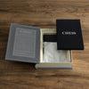 The Chess Vintage Bookshelf Edition sits open on a wooden table, displaying a cloth bag and "CHESS" booklet inside. Elegant gray and black packaging highlights the classic strategy game's timeless appeal.