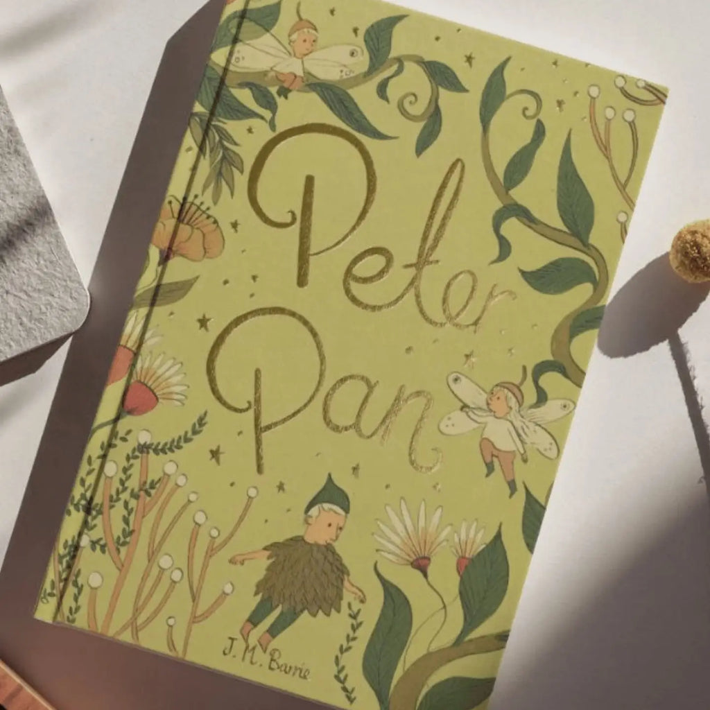 The Peter Pan | Barrie | Collector's Edition | Hardcover Book features an illustrated cover with fairies, plants, and whimsical green and gold designs evoking Never-Never Land’s magic under soft sunlight.