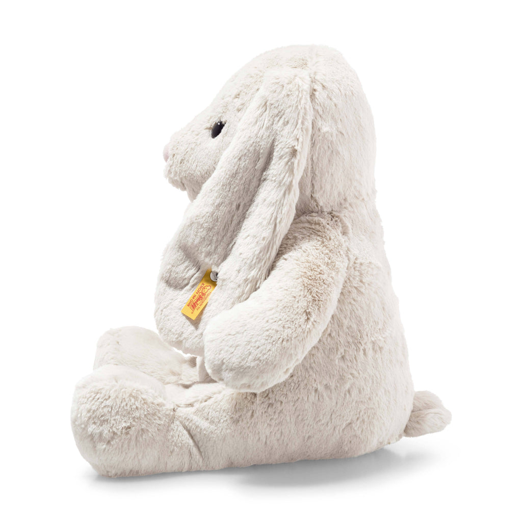 The Steiff Hoppie Bunny Stuffed Animal sits upright in profile, featuring long floppy ears, a small tail, and a yellow tag attached to one ear.