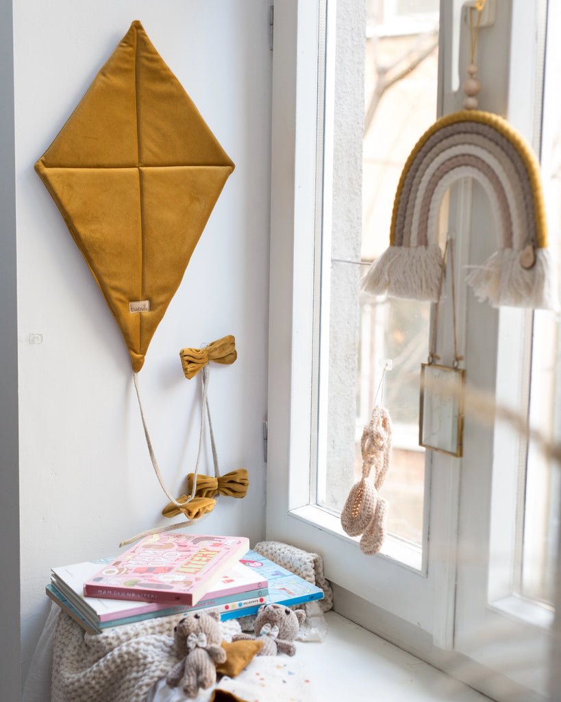 A cozy corner features a Velvet Kite Wall Decor in mustard, which is OekoTex®Standard100 certified, with bows hanging on the wall next to a white-framed window. Below, there is a stack of children's books, small knitted toys, and a soft blanket. A rainbow-shaped wall hanging and a stuffed animal decorate the children's room decor.