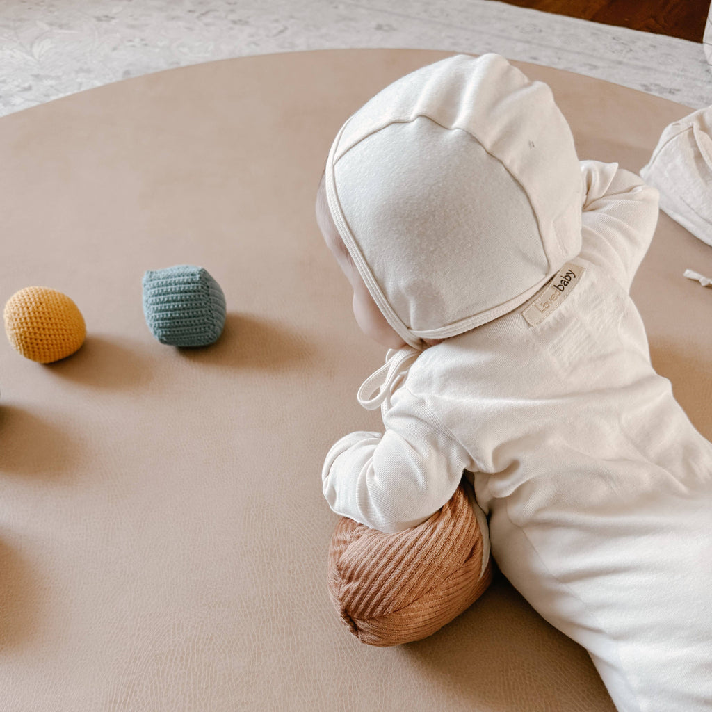 A baby in a cream-colored hooded outfit lies on a tan mat, propped up on the Tummy Time Baguette pillow, gazing at three textured toy blocks in yellow, blue, and green.