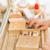 A child's hand plays with a Wooden Beauty Kit on a wicker tray, featuring lipsticks, compacts, and a handheld mirror. Painted in soft pastel colors with non-toxic paint, this set reflects the charm of a whimsical wooden vanity.