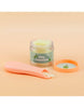 A small jar labeled "Tiny Tummies- Banana Jelly food - Tiny Harlow" is open with a beige-colored jelly inside. Next to it is an orange lid featuring a simple apple icon and a peach-colored baby spoon, which seems like the perfect magic spoon for your little one. The background features a soft peach shade.