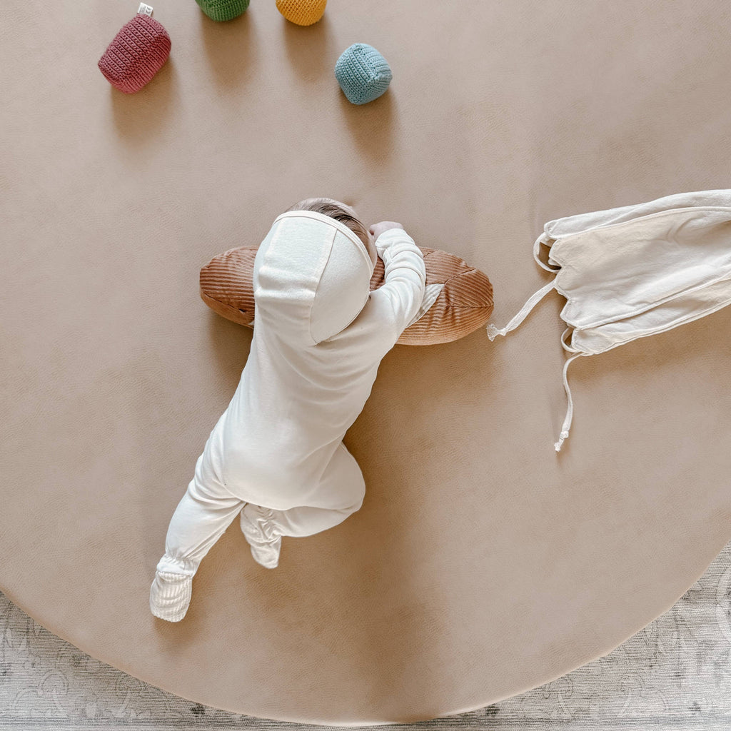 A baby in a white hooded onesie lies on a beige surface, reaching for the Tummy Time Baguette pillow. Colorful sensory beads and a white bonnet are nearby. The scene is viewed from above.