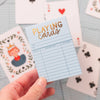 A hand holds the Sky Blue Library Card Deck Of Playing Cards, featuring gold and black text on a blue box. Several cards are spread out on a table in the background. Ships in approximately one week.