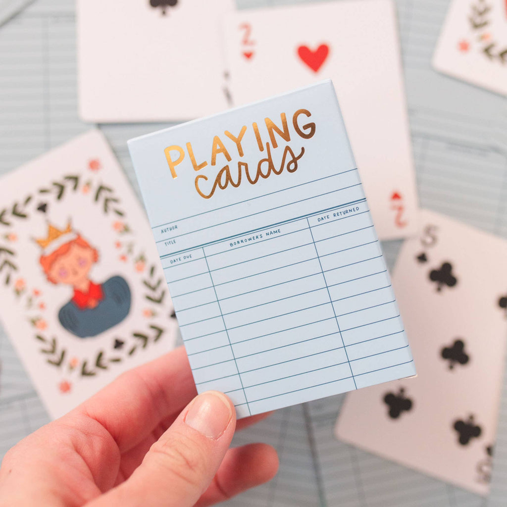 A hand holds the Sky Blue Library Card Deck Of Playing Cards, featuring gold and black text on a blue box. Several cards are spread out on a table in the background. Ships in approximately one week.