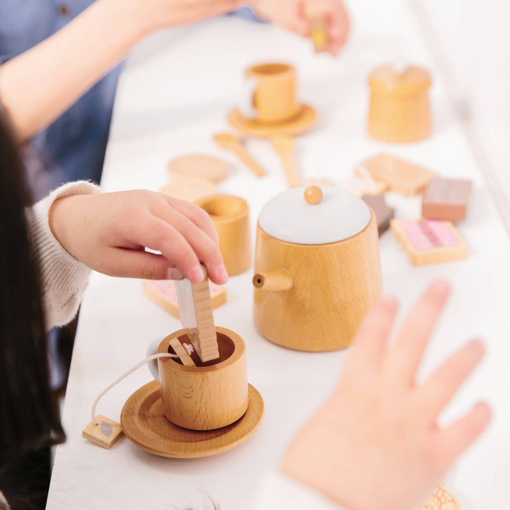 Children joyfully play with the Wooden Tea Set, dipping blocks into cups. The vibrant scene, complete with cups, a teapot, and wooden pieces, resembles a designer coffee table book showcasing imaginative play.