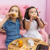 Two children play with a Wooden Tea Set on a pink couch, pretending to sip organic Matcha Tea from wooden cups. A table holds the tea set, honey, and waffles. One wears a blue dress, the other white. The light pink wall serves as their backdrop.