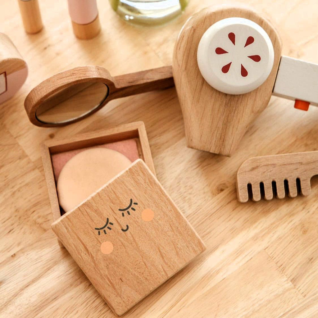 The Wooden Beauty Kit, a timeless toy set on a light wooden surface, includes a compact with a smiling face, makeup brush, comb, and beauty tools. It features natural wood finishes with minimal non-toxic paint details.