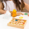 A child in a white dress with long brown hair joyfully plays in a vibrant space, arranging toy food on a table. Amidst the milk carton and honey bear bottle, the Wooden Tea Set for brewing organic Matcha Tea adds elegance as she explores the colorful wooden crate.