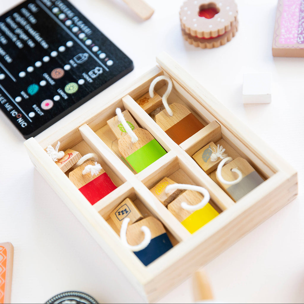 A wooden box with six compartments contains colored pegs and blocks in red, green, and blue hues. Nearby, a playful Wooden Tea Set mingles with various wooden toys and a black card on the crisp white play space surface adorned with symbols.