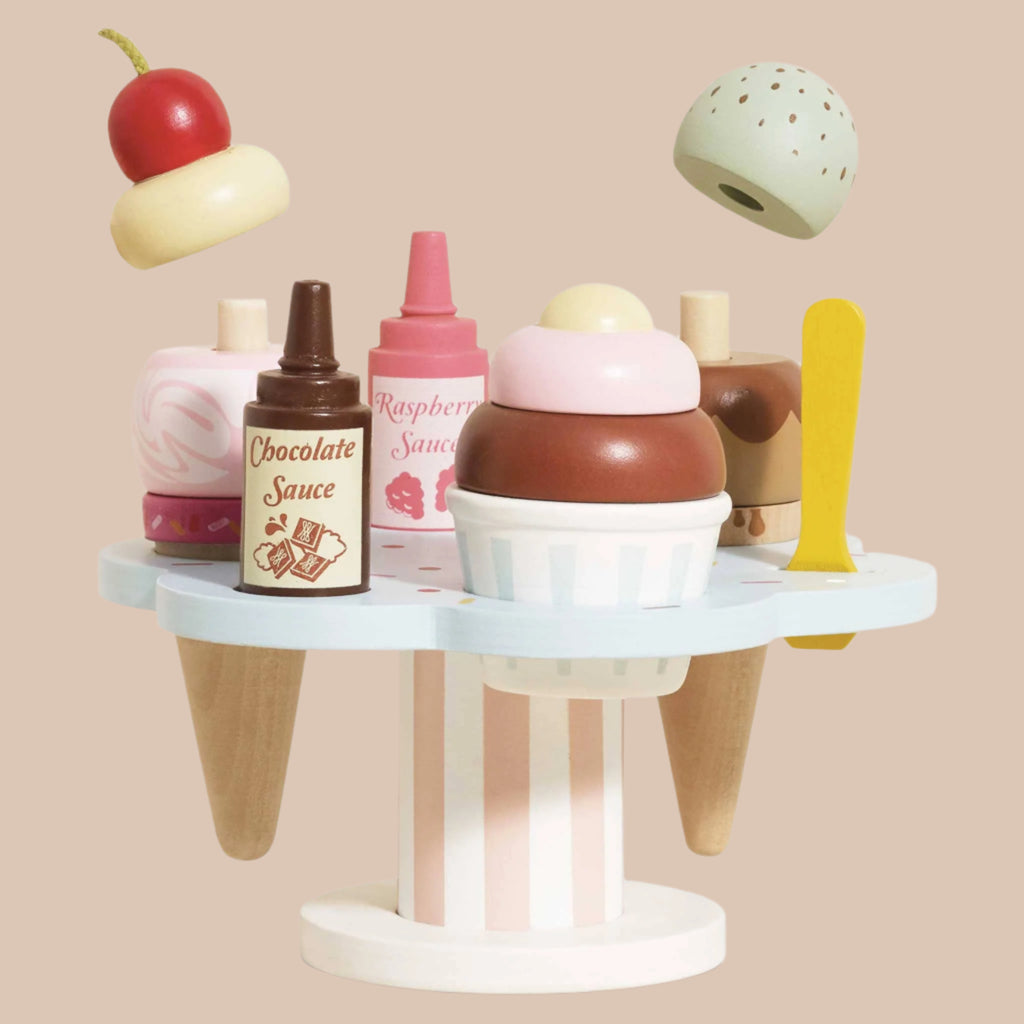The Wooden Ice Cream Stand & Toppings set showcases a light blue display with cones, interchangeable scoops, toppings, and chocolate and raspberry sauce bottles. A red cherry and green scoop are suspended above on a beige backdrop, inviting role-play fun.