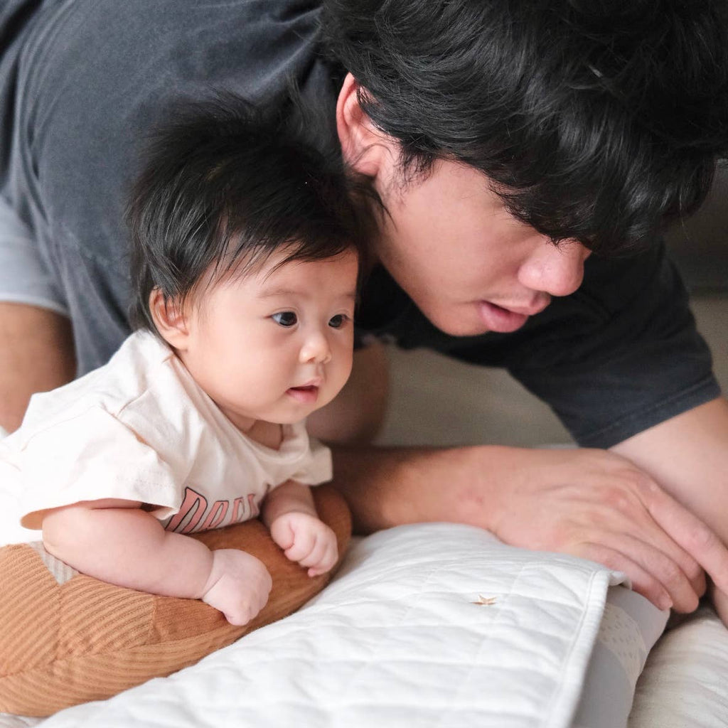 A baby with dark hair lies on their stomach on a bed, propped up on a Tummy Time Baguette pillow, while a dark-haired adult leans close, both focused intently on something out of frame.
