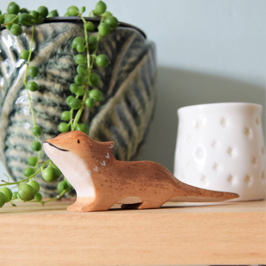 A small Bearded Dragon wooden animal, crafted from sustainable hardwood, sits on a wooden surface. Behind it, a plant in a decorative pot with leaf patterns stands alongside a white candle holder adorned with star shapes.