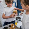 Two children play with the Wooden Tea Set, one holding a cup while the other pours. In their rustic room with mottled walls and small stools, designer coffee table books add charm to the playful scene.