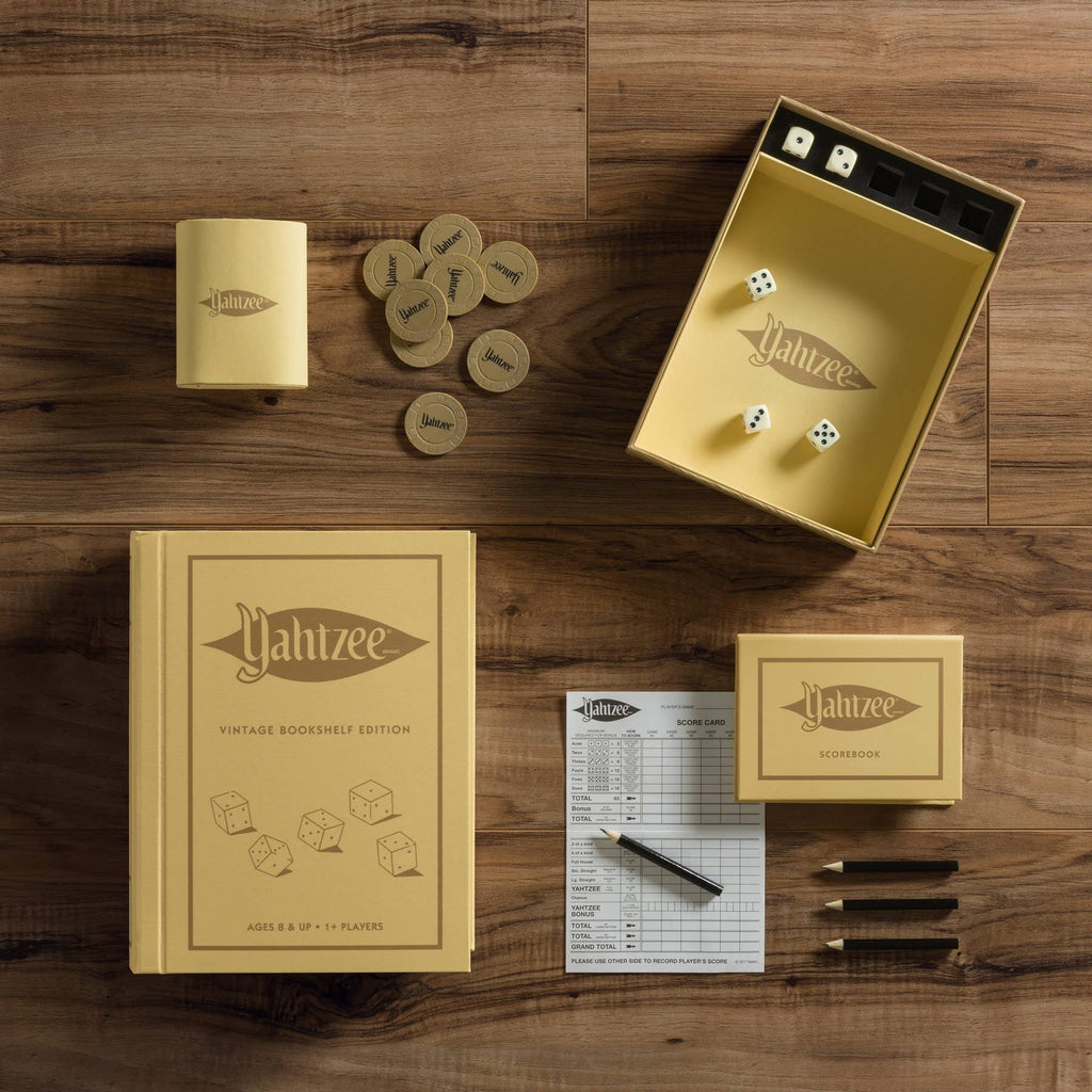 The Yahtzee Vintage Bookshelf Edition, displayed on a wooden surface, features a book-shaped box, dice, scorepad, pencils, chips, and a dice cup—all in vintage yellow and brown tones.