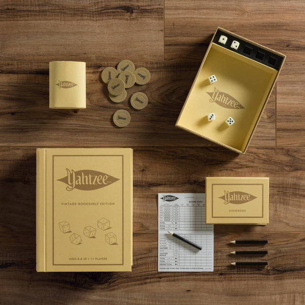 The Yahtzee Vintage Bookshelf Edition, displayed on a wooden surface, features a book-shaped box, dice, scorepad, pencils, chips, and a dice cup—all in vintage yellow and brown tones.